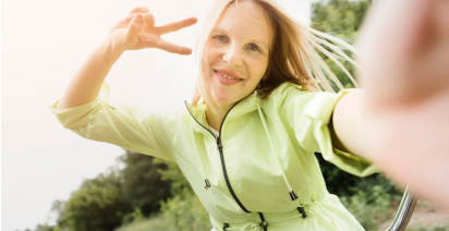 Smiling blonde woman taking a selfie outdoors in a light green jacket, holding up a peace sign.