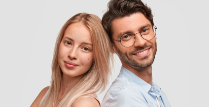 A smiling young man wearing round glasses leaning back-to-back with a blonde woman against a light background.