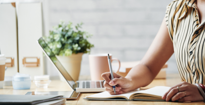Woman writing in a notebook at a bright desk next to an open laptop and a potted plant.