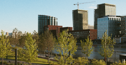 A leafy waterfront park with modern city high-rises and a construction crane in the background under a clear sky.
