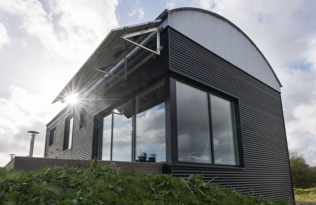 Modern home with black corrugated metal cladding, a curved barrel roof, large glass windows, and roof-mounted solar panels.