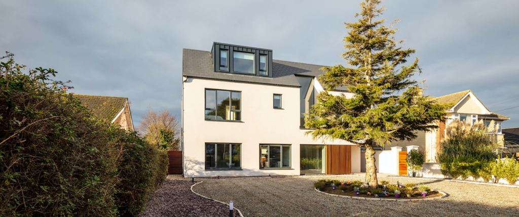 Modern three-story white home with dark roof dormers, a gravel driveway, and a central evergreen tree.