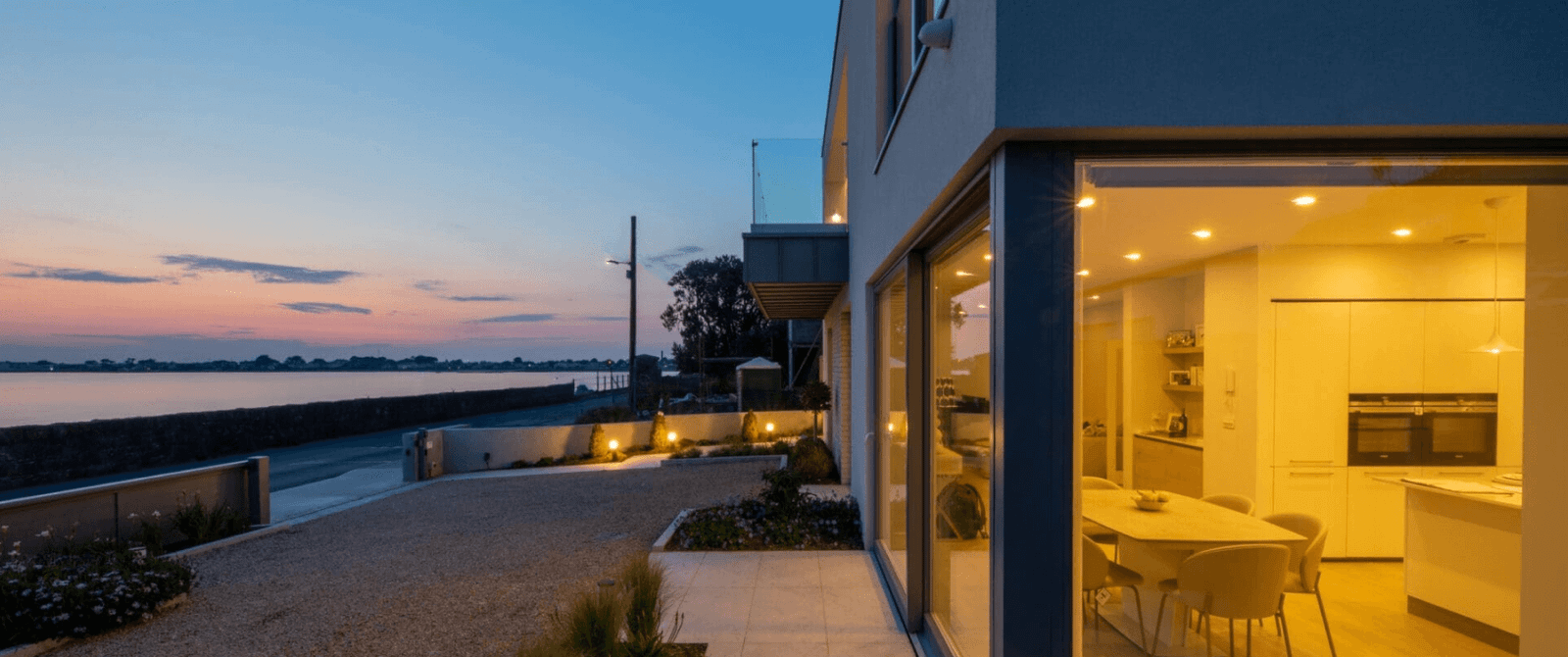 Modern waterfront home with illuminated interiors, overlooking a scenic ocean bay at twilight.