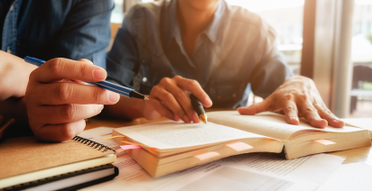 Two people studying together at a table, holding pens and reviewing an open textbook filled with sticky note markers.