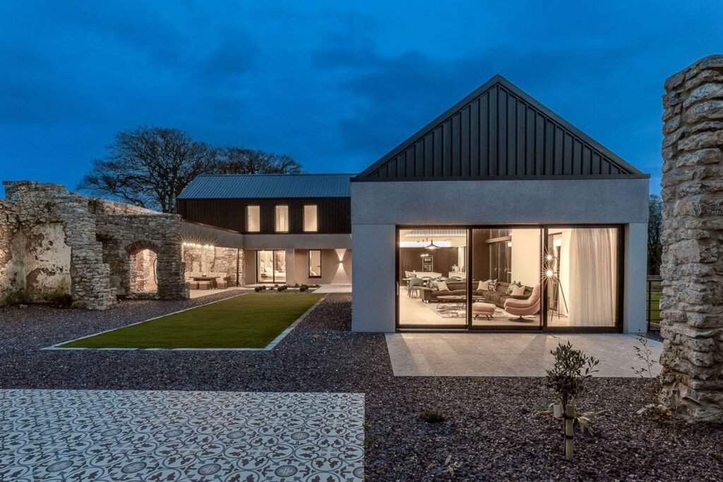 Modern home at dusk featuring glass sliding doors, lit interior spaces, and historic stone ruin walls built into the courtyard design.