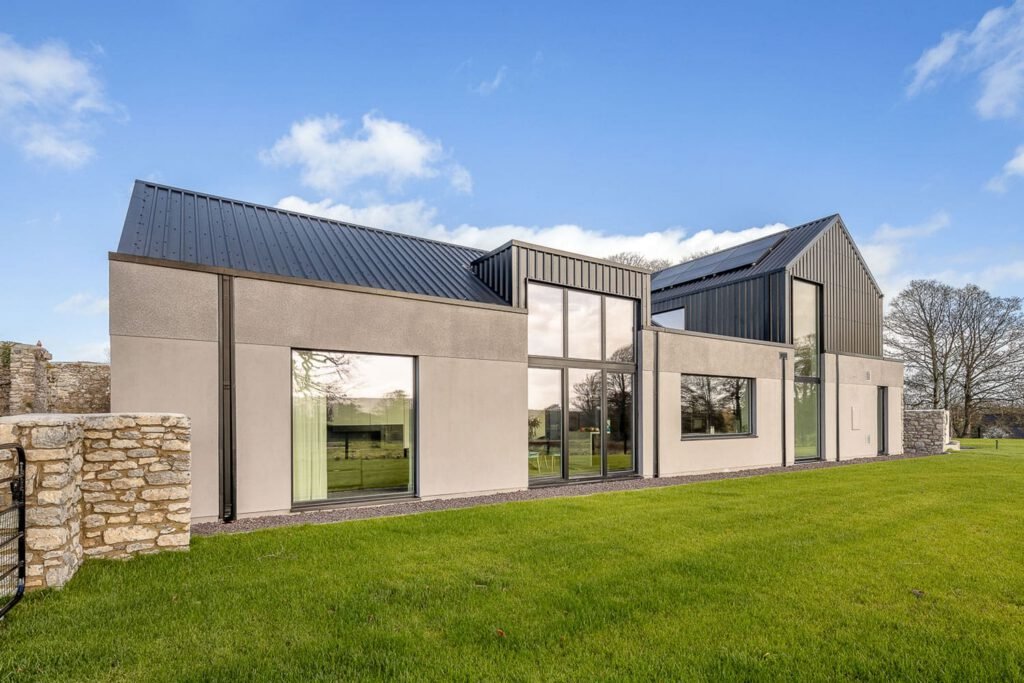 Modern home with grey walls, dark metal roof, large glass windows, and a grassy lawn next to a stone wall.