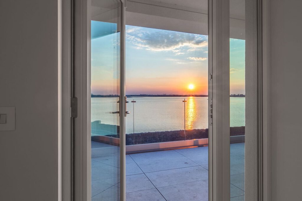 View through open white glass doors of a balcony overlooking a lake sunset.