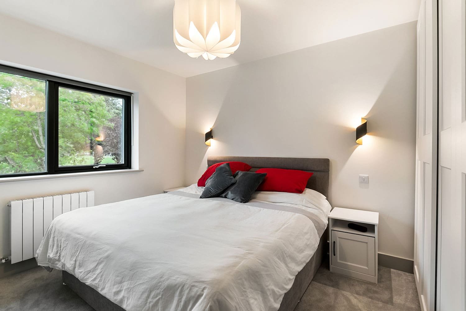 Modern bedroom with a double bed, red and grey pillows, wall sconces, and a flower-shaped ceiling light.