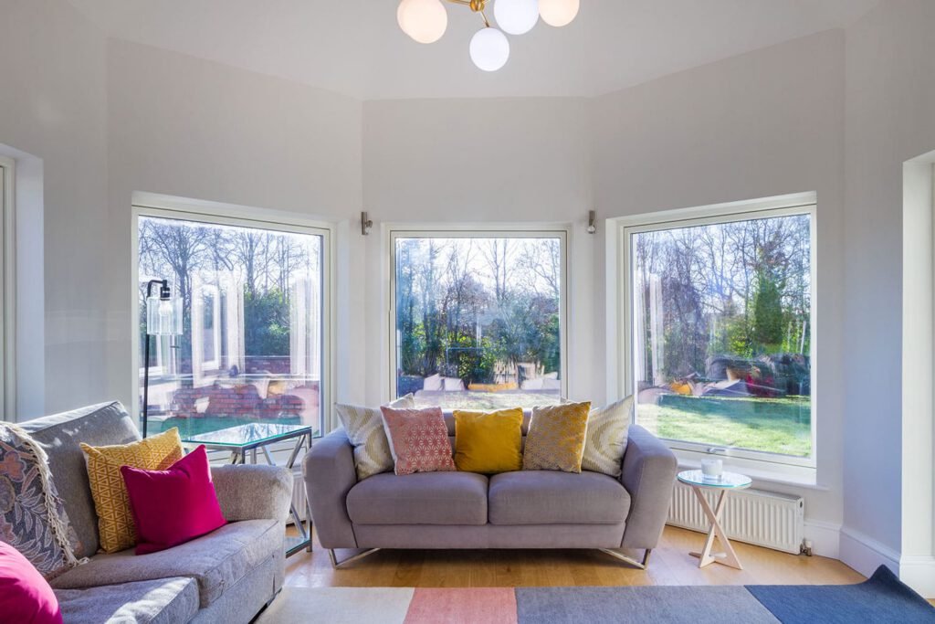 Sunlit living room featuring grey sofas with colorful accent pillows in front of large bay windows.