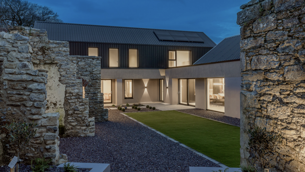 Modern house built alongside ancient stone ruins with a gravel courtyard at dusk.