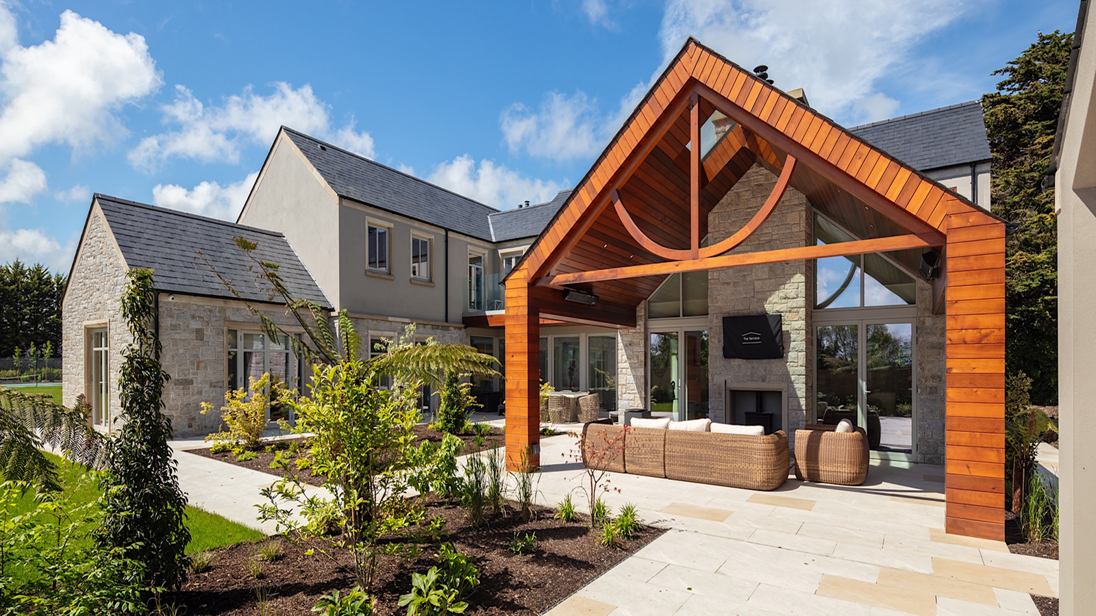 Modern house with a large wooden covered patio, outdoor furniture, and a stone fireplace.