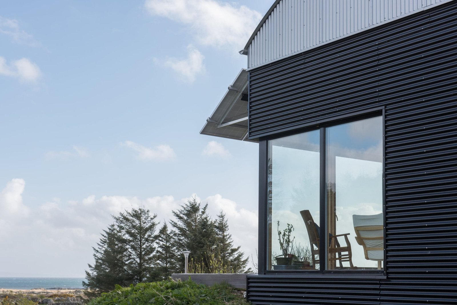Modern home with black corrugated metal cladding, a curved barrel roof, large glass windows, and roof-mounted solar panels.