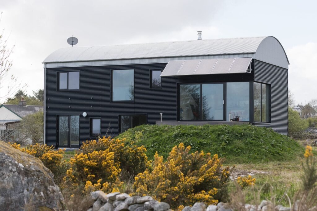 Modern home with black corrugated metal cladding, a curved barrel roof, large glass windows, and roof-mounted solar panels.
