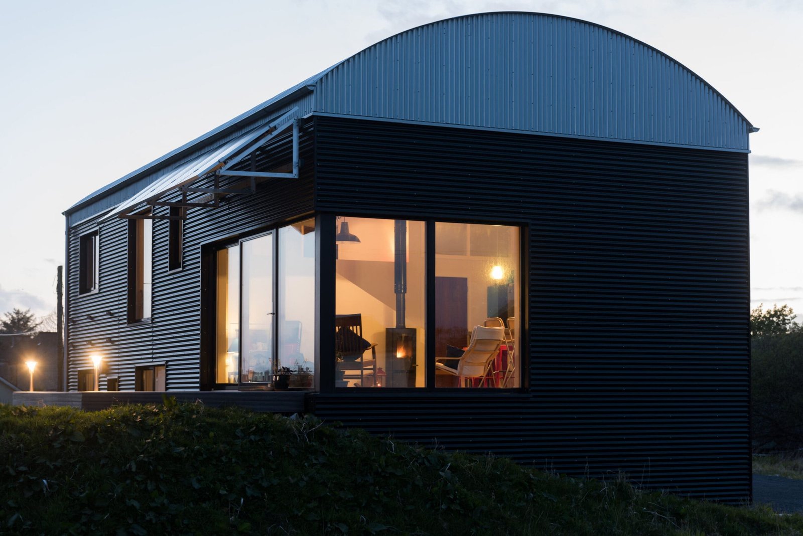 A modern house with dark corrugated metal siding, solar panels, and a curved roof under a sunny sky.