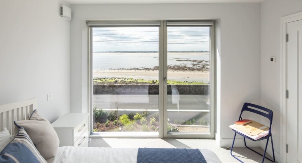Bright bedroom with light grey walls, carpeted floor, and large glass door with a sea view.