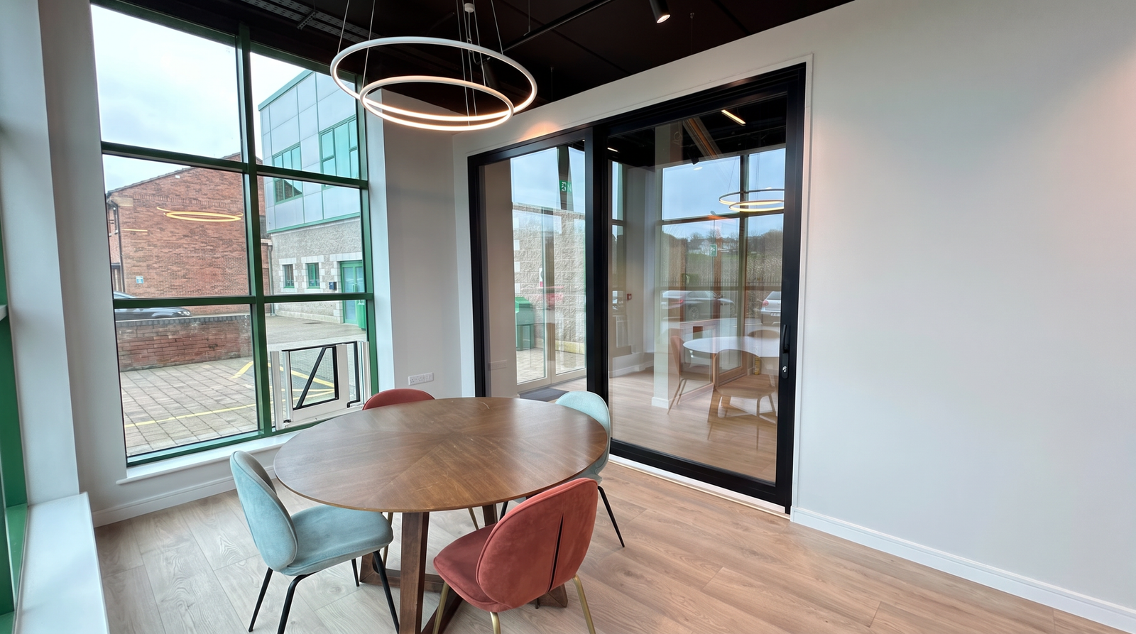 Modern office meeting space with a round wooden table, colorful chairs, and large glass windows.
