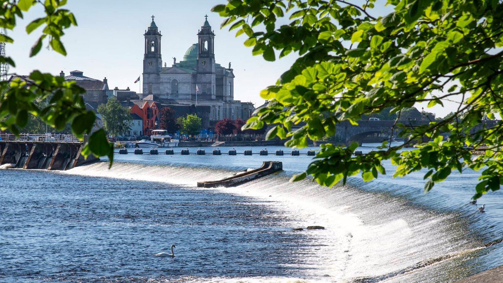 Athlone Castle and St. Peter and Paul's Church overlooking a weir on the River Shannon, framed by green leaves on a bright sunny day.