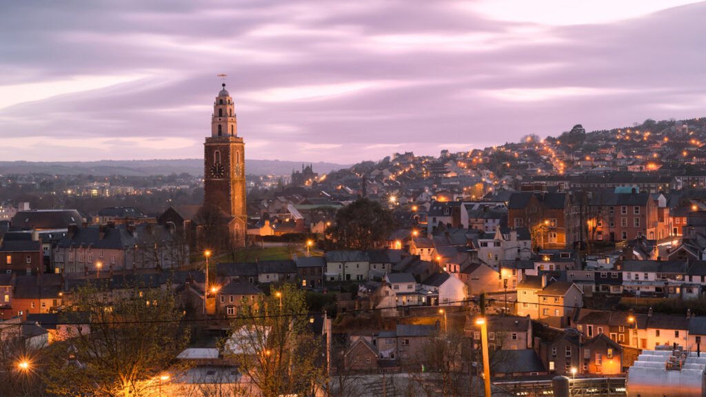 Shandon Steeple rising above the illuminated city landscape of Cork, Ireland, under a purple twilight sky.