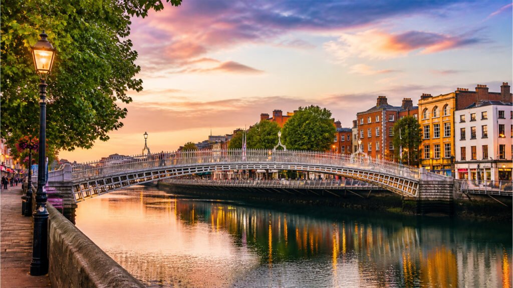 The Ha'penny Bridge spanning the River Liffey in Dublin under a colorful sunset sky, flanked by streetlamps and historic buildings.