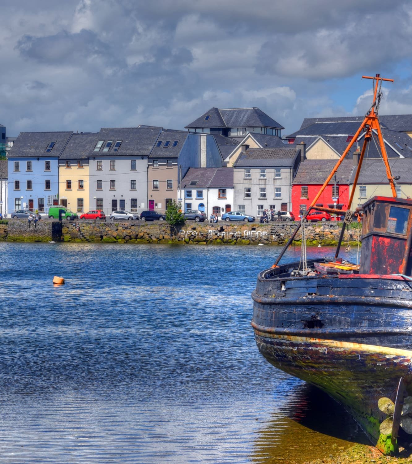 Colorful waterfront houses along Claddagh Quay in Galway, with a rustic fishing boat moored in the foreground.