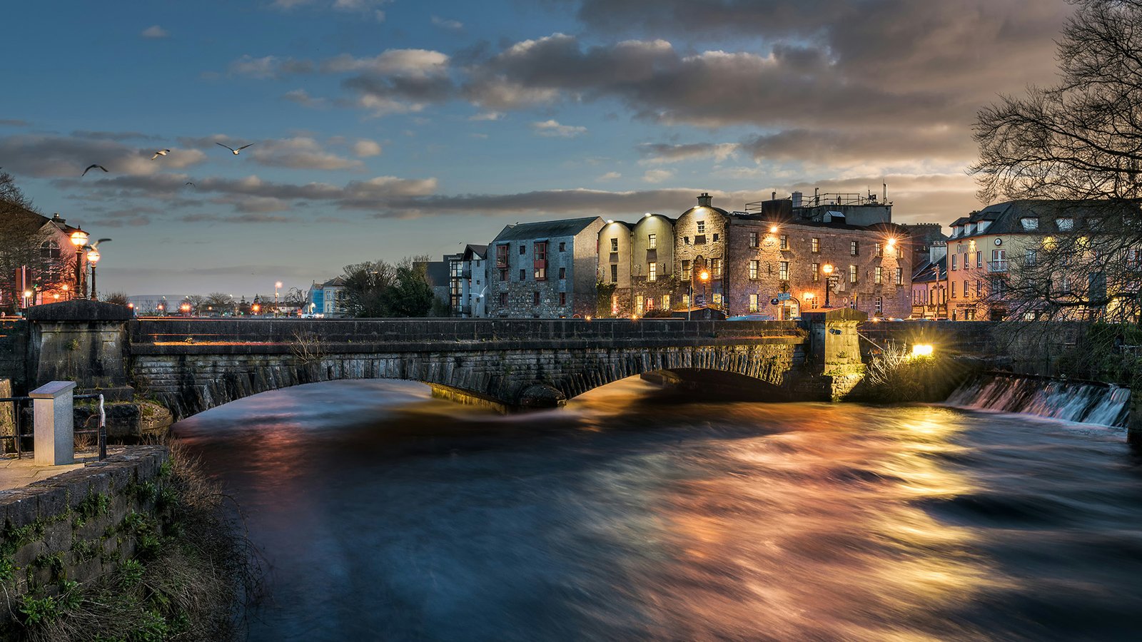 A stone arch bridge spanning the River Corrib in Galway at dusk, with illuminated historic buildings reflected in the water.