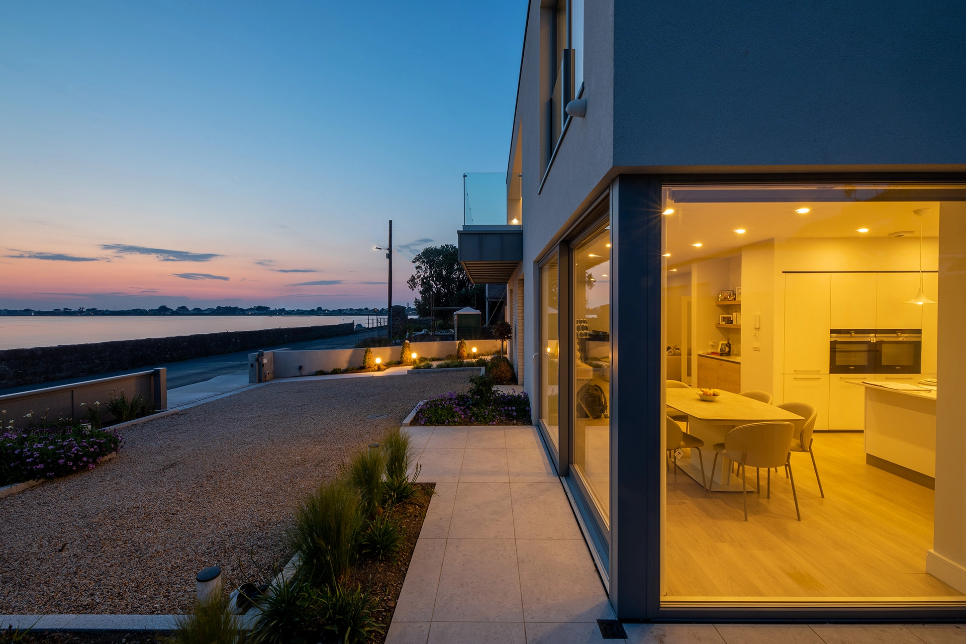 Modern waterfront home exterior illuminated at sunset with large glass windows showing the dining area.