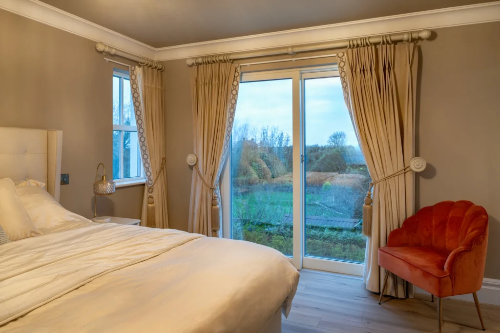 Elegant bedroom with beige curtains, double bed, rust velvet armchair, and sliding glass patio doors.