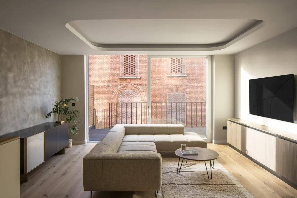 Modern living room with modular beige sofa, round coffee table, and large glass door opening to a terrace facing a brick building.