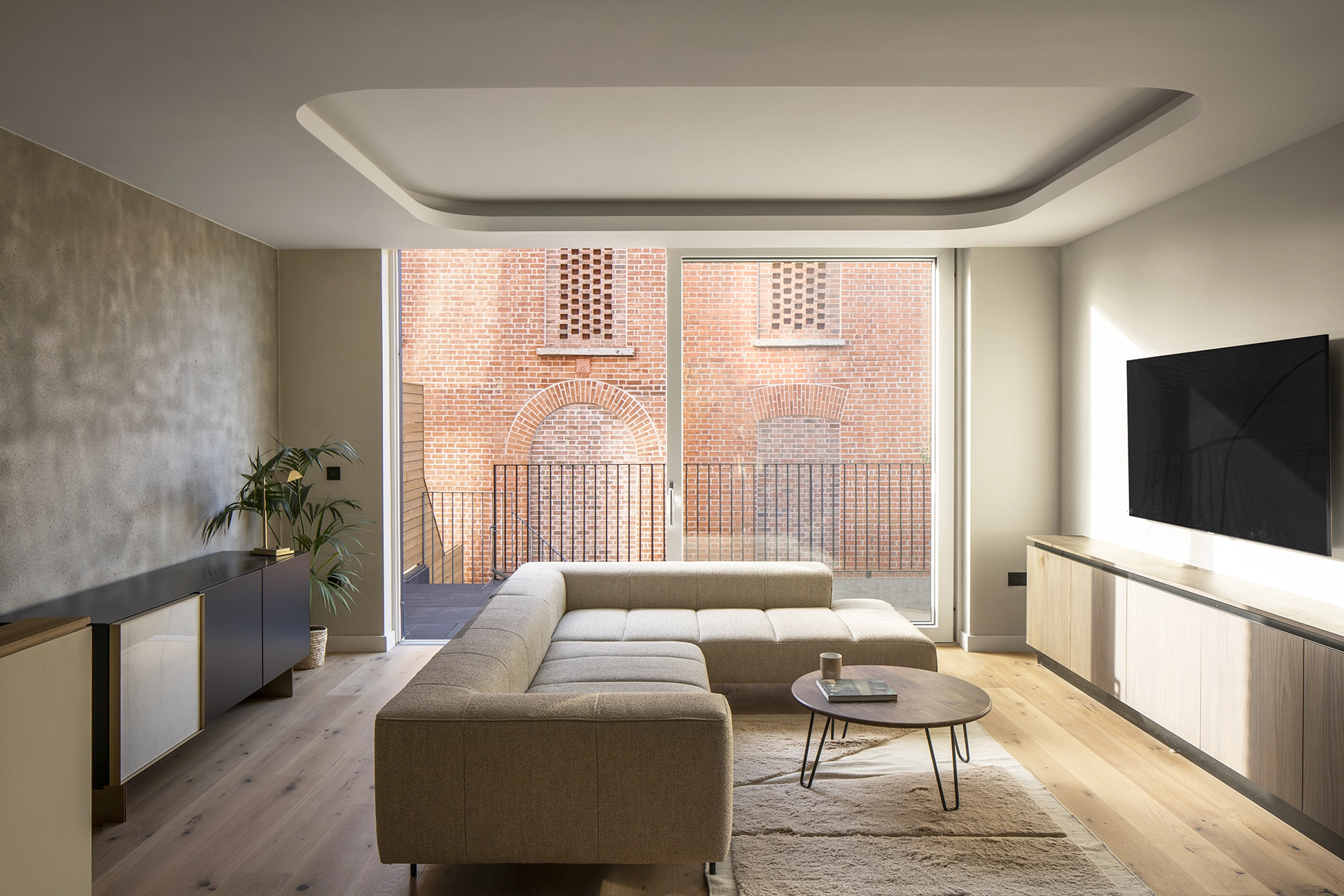 Modern living room with modular beige sofa, round coffee table, and large glass door opening to a terrace facing a brick building.